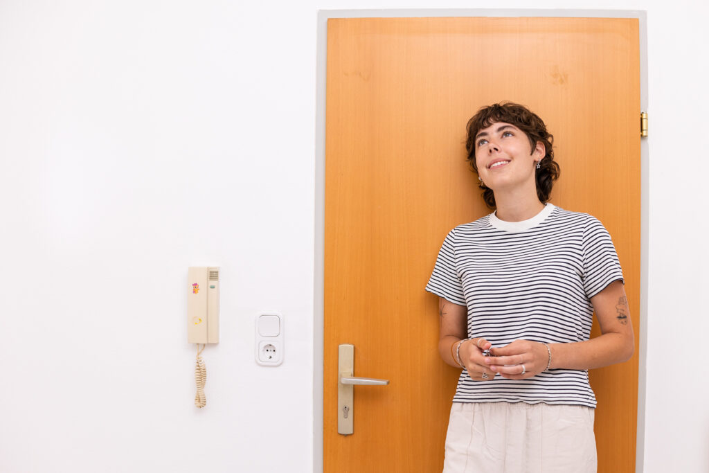 Portraitfoto von Paula. Sie lehnt glücklich an der Türe mit dem Schlüssel in der Hand.