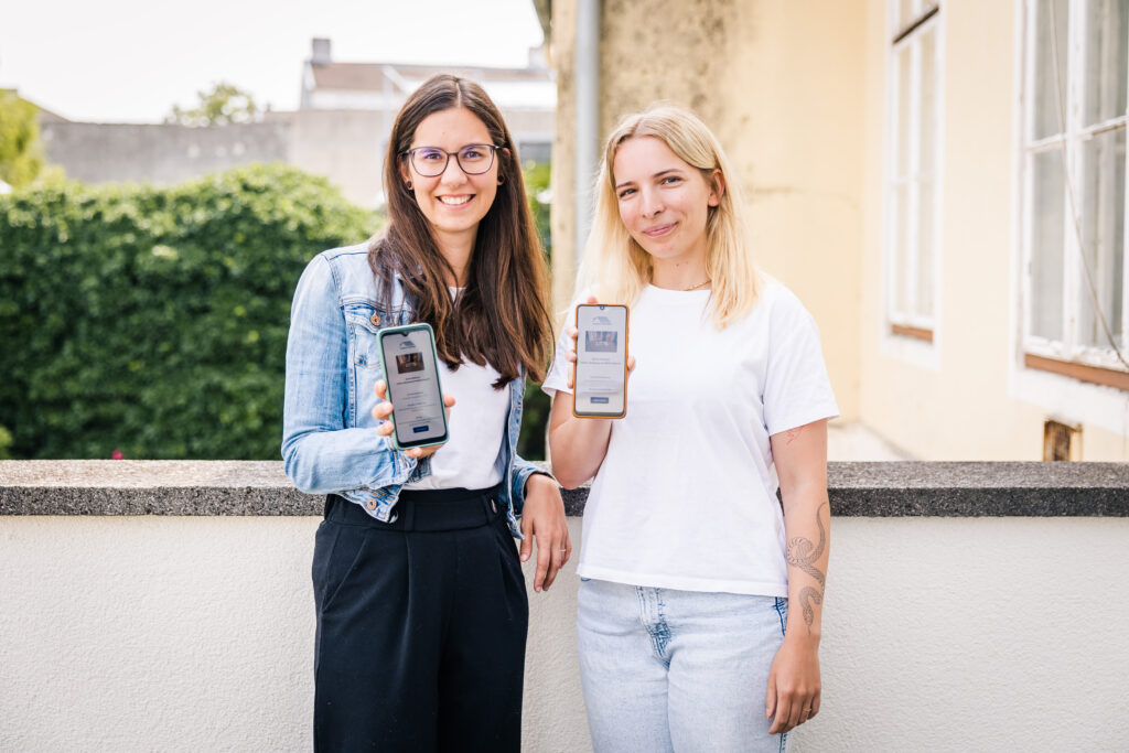Zwei Mitarbeiterinnen vom Verein Wohnen zeigen Smartphone mit App in die Kamera.